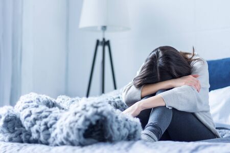Young woman in gray clothes is sitting curled up on bed at home. Upset brunette girl is suffering, crying. Depression because of loneliness, stress, problems. Psychological disorders concept.の写真素材