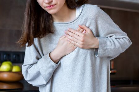 Young woman in grey clothes is holding hands on breast. Brunette girl is feeling bad. Sudden heart attack, myocardial infarction at home. Effect of stress and unhealthy lifestyle concept.の写真素材