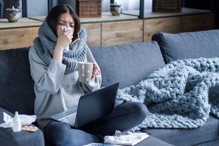 Ill brunette girl is sitting at sofa and working remotely on laptop. Female is blowing out snot, having fever and headache. Young woman is treated at home, took sick leave. Winter cold and flu conceptの写真素材