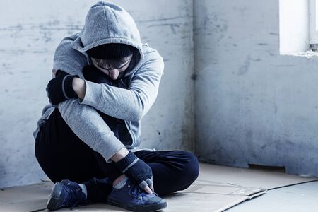 Unhappy guy in hood is sitting on cardboard on floor in abandoned building. Homeless sad man lost everything because of addiction. Social problems in society. Loneliness and depression concept.の写真素材