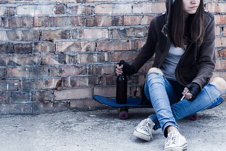 Young woman in hood is sitting on skateboard and holding smoldering cigarette and bottle of beer. Homeless teenage girl is drinking and smoking in abandoned building. Dregs of society concept.の写真素材