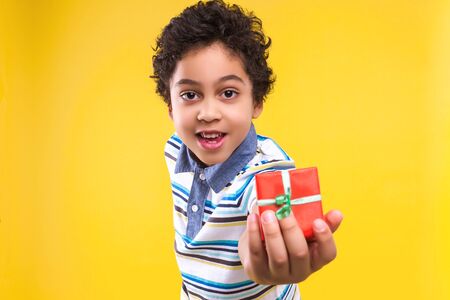 Curly smiling dark skinned boy is holding gift box, wrapped on red paper and green ribbon. Black child dressed in striped blue t-shirt is giving present. Happiness and surprise concept.の写真素材