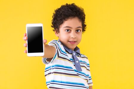 Curly pretty dark skinned boy is holding in hands white smartphone with empty blank screen display....