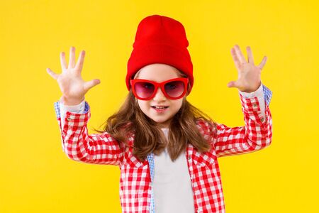 Beautiful cheerful stylish little girl dressed in pink checkered shirt, red cap and jeans on yellow background. Pretty cute child is showing with her hands number ten. Emotional portrait concept.の写真素材