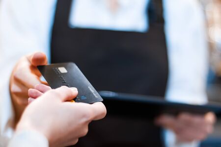 Closeup woman barista, waiter in brown apron uniform is holding tablet computer, taking credit card for order from client customer in cafe, restaurant. Visitor service. Cashless payments concept.の写真素材