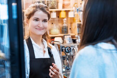 Young woman barista in brown apron is taking order for preparing espresso americano from client in...