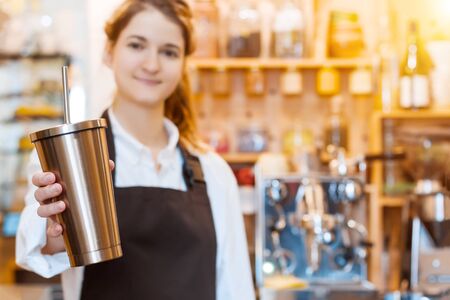 Closeup female hands are holding stainless metal reusable tumbler cup mug with drinking straw. Barista woman  prepared, brewed coffee using professional machine in cafe, restaurant. Take away, to go.の写真素材