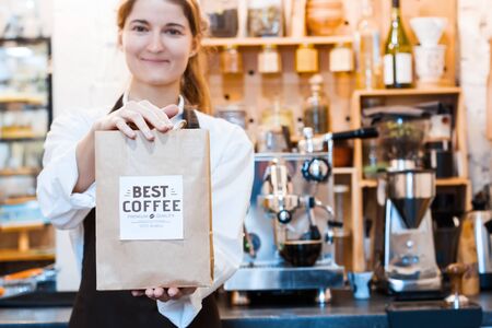 Young woman barista in brown apron is holding craft packet with arabica beans, ready to brew coffee to client customer in cafe, restaurant. Professional machine for preparing americano, espresso.の写真素材