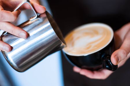 Closeup of female hands holding metal jug with hot milk and froth. Process of preparing, brewing americano, latte, capuchino. Barista woman is making coffee in cafe. Hot drinks, beverages concept.の写真素材