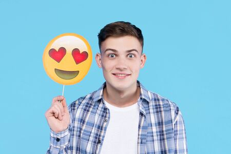 Young man in white t-shirt, checkered shirt is holding in hand paper decorative emoticon, emoji icon, sign. Guy student is posing with photo props on blue background. Emotional portrait concept.の写真素材