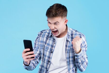 Satisfied, happy, wondered student guy is holding in hand smartphone, looking at screen display. Young man dressed in checkered shirt, white t-shirt on blue background. Modern devices concept.の写真素材