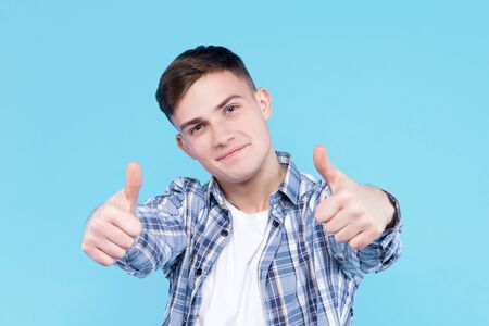 Satisfied pleased guy dressed in white t-shirt, checkered shirt is standing on blue background. Young man is showing positive thumbs up sign. Male gesturing. Emotional portrait concept.の写真素材