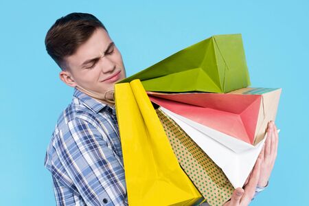 Young tired man in checkered shirt is holding lot of colorful paper bags, packages, packets on blue...