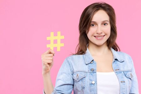Pretty girl user of social media in white t-shirt, denim jacket is holding in hand yellow paper hashtag sign. Young woman blogger is smiling on pink background. Emotional portrait concept.の写真素材