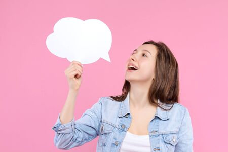 Pretty girl in white t-shirt, denim jacket is holding in hand paper decorative thinking, speaking cloud. Young woman is smiling and dreaming on pink background. Emotional portrait concept.の写真素材
