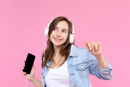 Pretty, smiling girl is holding in hand smartphone and listening music. Young woman dressed in denim...