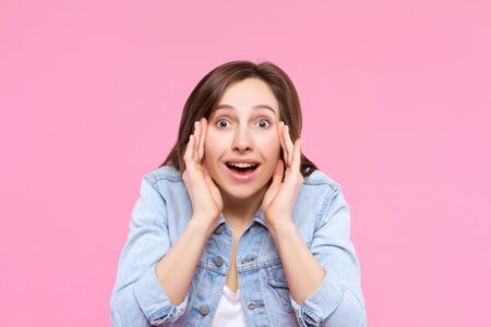 Pretty girl dressed in white t-shirt, denim jacket is standing on pink background. Young surprised...
