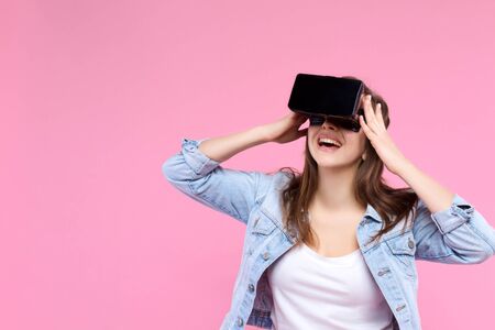 Pretty, smiling, surprised girl in VR glasses on pink background. Young woman dressed in denim jacket, white t-shirt is enjoying virtual reality world. Modern technology and devices concept.の写真素材