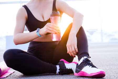Closeup young athletic woman in black tracksuit is sitting outdoor at sunset, holding pink bottle of water. Girl is relaxing after training, stretching, yoga. Daily routine. Sport lifestyle.の写真素材