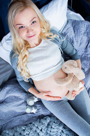 Pregnant beautiful woman waiting for a child holding toy rabbit close to beautiful belly, tummy. Laying on gray bed sheets next to cute baby socks. Preparation for childbirth maternity parents conceptの写真素材