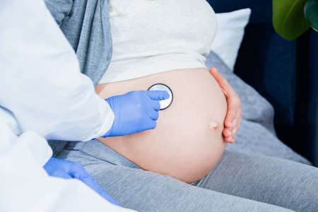 Closeup of big pregnant female belly, tummy. Doctor is listening sounds in abdomen with medical...