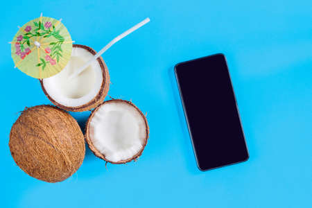 Smartphone with blank screen, halves of coconut nut, milk cocktail are laid out on blue background. Tourism application for buying airline tickets, tours to foreign tropical exotic countries concept.の写真素材