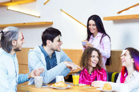 Young people guys and girls are having meeting, sitting at table in cafe. Cheerful students friends are playing games with stickers, rejoicing, spending time together. Leisure and rest mood.の写真素材