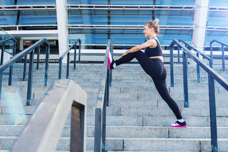 Young athletic woman in black tracksuit uniform is training outdoor on street. Blonde girl is...