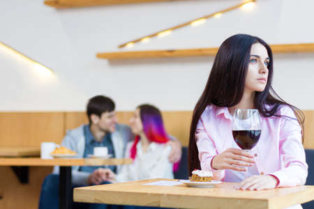 Lonely sad girl is sitting alone at table in cafe, restaurant. Young woman without relationships is spending time with herself. Loving couple is on date on background. Loneliness concept.の写真素材