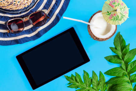 Tablet computer, striped hat, sun glasses, palm leaves, coconut halves, cocktail are laid out on blue background. Tourism application for buying airline tickets, tours to foreign countries concept.の写真素材