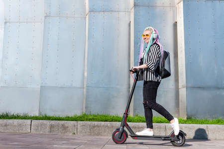 Smiling girl with colorful dreadlocks is riding, driving electric scooter in street of modern city....