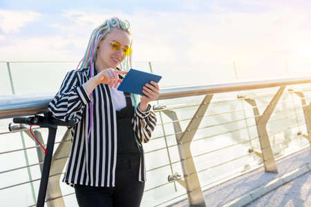 Girl with colored dreadlocks is standing on bridge, holding tablet. Young woman in yellow glasses...