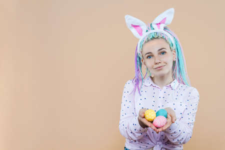 Pretty girl in bunny ears, pink shirt is holding eggs. Young woman with colored dreadlocks is preparing for celebration. Happy easter, spring concept. Carnival, seasonal party decor for holiday.の写真素材