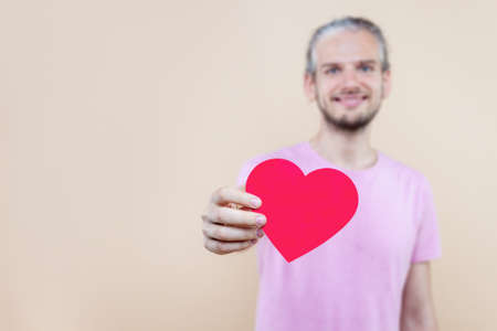 Cute smiling boy is holding red decorative sign of heart. Young man cupid in pink t-shirt is expressing sympathy, feelings. Happy St. Valentine's Day. Fall in love. Like concept.の写真素材