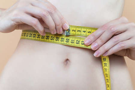 Closeup of female slender tummy tied with yellow centimeter tape. Young woman is measuring parameters of figure. Diet nutrition, weight loss concept. Healthy, sports lifestyle. Disease of anorexia.の写真素材