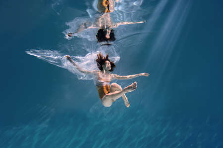 Fashionable and athletic girl free diver alone in the depths of the ocean. Swimmer brunette diving deep in ocean on blue underwater background. Pollution, plastic and ecology conceptの写真素材