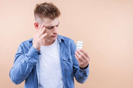 Young handsome man in denim shirt standing over beige background tired rubbing, touching head, feeling fatigue, headache. Male is holding pain pills. Stress and frustration concept. Cold flu symptoms.の写真素材