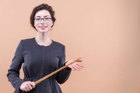 Portrait of teacher keeping wooden pointer showing blank space, smiling isolated on pastel beige color background. Young woman lecturer in casual jacket is ready to teach students. Study at universityの写真素材