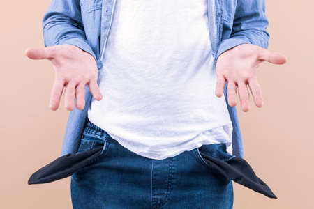Closeup man is holding his turning out empty pants jeans pocket to show no cash inside isolated on beige background. Unemployed male spent money. Financial crisis, problems, bankruptcy, poverty.の写真素材