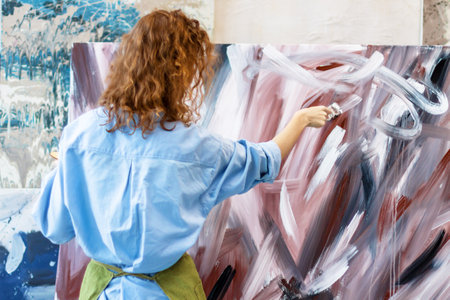 Student girl of faculty of arts wearing blue t-shirt standing in front of easel, mixing colors using paint brush while working on painting in university studio. Inspiration and creativity conceptの写真素材
