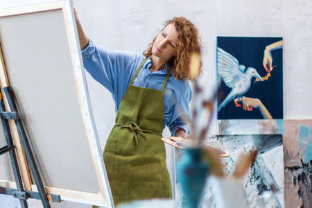 Woman artist is drawing on canvas with brush, palette, in workshop studio gallery. Process of making...