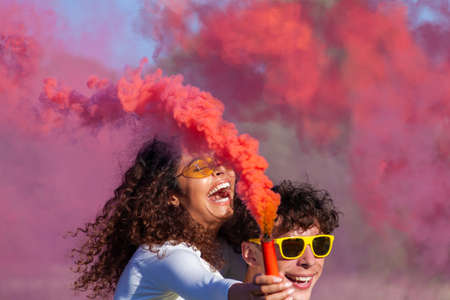 Beautiful young man and woman hold light up colored smoke bombs - Happy friends having fun in the park with multicolored smoke bombs - Young students celebrating spring break together. Holi festival.の写真素材