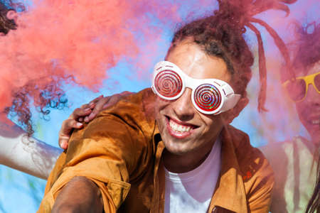 Beautiful young man hold light up colored smoke bombs - Happy friends having fun in the park with multicolored fog bombs - Young students celebrating spring break together. Holi festival concept.の写真素材