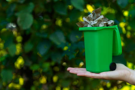 Female hand is holding green plastic recycle garbage bin at park. Waste recycling sorting management. Reduce and reuse trash concept. Protection of environment from pollution. Earth day. Save planet.の写真素材