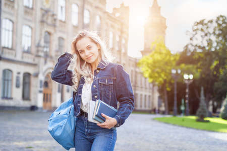 Attractive female student with backpack walking to campus, education concept. Blonde girl is smiling and holding books in her hands on college background. Young woman is taking exams at university.の写真素材