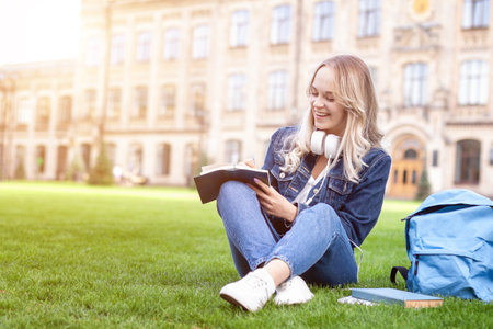 Blond female student is sitting on green grass yard near university campus. Young woman is studying and listening music with headphones outdoor. Back to high school concept. Education at college.の写真素材