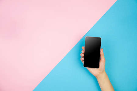 Young woman hand holding smartphone with black screen on light pink blue table background. Copy space empty place for text inscription. Mobile application interface concept design. Modern technology.の写真素材