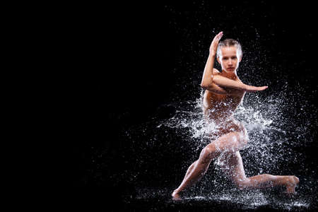 Wet young woman in tight beige swimsuit is dancing on floor under rain and splashes, drops of water. Girl modern dancer is doing tricks. Contemporary art dance. Freedom and freshness concept.の写真素材