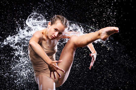 Wet young woman in tight beige swimsuit is dancing on floor under rain and splashes, drops of water. Girl modern dancer is doing tricks. Contemporary art dance. Freedom and freshness concept.の写真素材