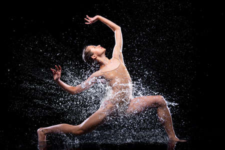 Wet young woman in tight beige swimsuit is dancing on floor under rain and splashes, drops of water....